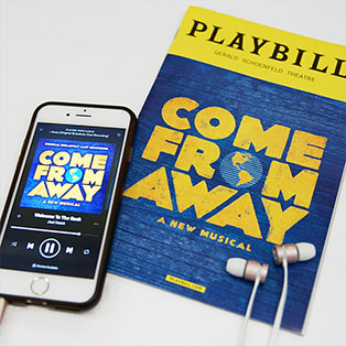 Thumbnail image of Come From Away’s Playbill and Come From Away album playing on smartphone with headphones